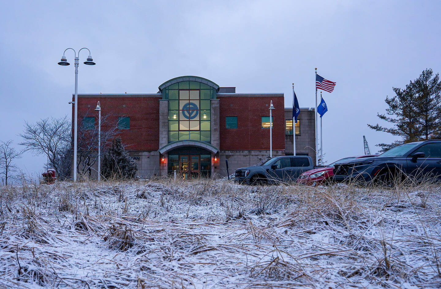 A brick building with a glass entrance and flags stands behind a snow-dusted field, with vehicles parked nearby under an overcast sky.